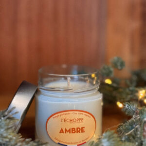 Bougie parfumée à l'ambre dans un pot en verre avec couvercle métal posé à côté, contre un mur en bois.