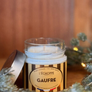 Bougie parfumée à la gaufre dans un pot en verre avec couvercle métal posé à côté, contre un mur en bois.