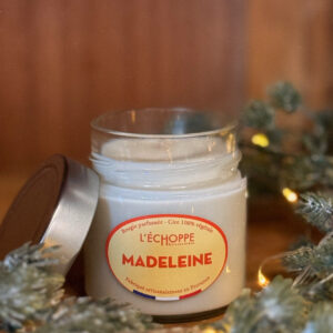 Bougie parfumée à la madeleine dans un pot en verre avec couvercle métal posé à côté, contre un mur en bois.