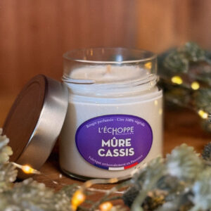 Bougie parfumée mûre cassis dans un pot en verre avec couvercle métal posé à côté, contre un mur en bois.