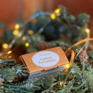 Diffuseur vanille patchouli dans son étui papier, posée au milieu d’une guirlande de sapin avec pommes de pin et petites lumières chaleureuses, ambiance décorative de Noël.