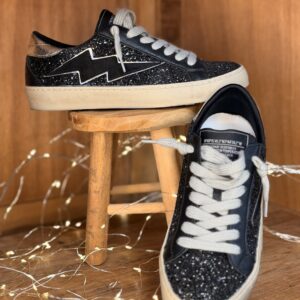Basket femme noire à paillettes avec éclair latéral noir, talon métallisé argent, lacets gris et semelle crème, posée sur tabouret en bois avec deuxième chaussure à l’arrière et guirlande lumineuse.