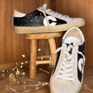 Basket femme noire à paillettes, empiècements cuir noir et blanc, logo rond blanc sur le côté, lacets blancs, présentée sur tabouret en bois avec seconde chaussure de profil derrière et guirlande lumineuse.