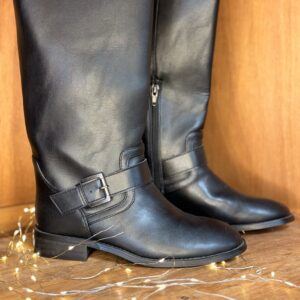 Bottes cavalières femme noir mat hauteur mi-mollet, bride à boucle sur cheville et zip côté intérieur, posées sur étagère en bois avec guirlande lumineuse dorée au sol