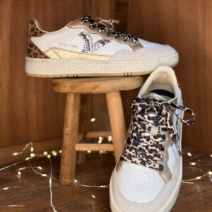 Basket femme Victoria blanche et beige avec empiècements dorés et imprimé léopard, lacets léopard, semelle crème, posée sur tabouret en bois avec deuxième chaussure de profil en arrière-plan et guirlande lumineuse