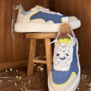 Basket femme blanche et beige avec empiècements denim bleu et touches jaune fluo, semelle épaisse blanche, présentée sur tabouret en bois avec seconde chaussure à l’arrière et guirlande lumineuse décorative