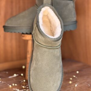 Bottines femme vert olive mouton synthétique, tige courte, semelle crantée noire, présentées sur tabouret en bois avec guirlande lumineuse.