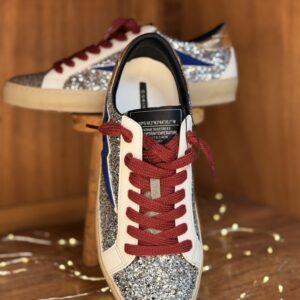 Basket femme argentée pailletée avec empiècements bleu marine, lacets rouges, logo discret, semelle blanche, posée sur tabouret en bois avec guirlande lumineuse