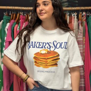 Une jeune femme brune aux cheveux longs pose avec sourire dans une boutique, portant un t-shirt blanc taille unique “Baker’s Soul” illustré de pancakes dorés superposés et du texte “Rising Process.,