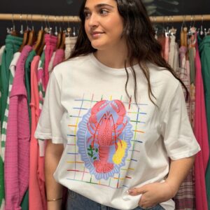 Une jeune femme brune aux cheveux longs pose avec sourire dans une boutique, portant un t-shirt blanc taille unique illustré d'un homard daux couleurs rose, bleu et jaune, vu de face.