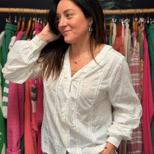 Jeune femme brune pose de face, une main dans les cheveux, portant une chemise en coton plumetis blanc à manches longues et col V, dans une boutique colorée. Look confortable et tendance.