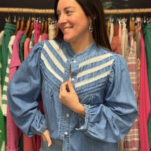 Jeune femme brune sourit de profil en boutique, portant chemise bleu denim à volants blancs guipure lavallière et manches bouffantes. Style romantique bohème chic.