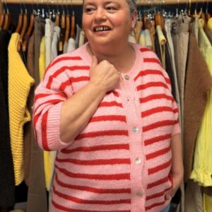 Femme ronde portant un gilet rayé rose et rouge en maille douce, boutonné sur le devant, posant dans une boutique de vêtements grande taille avec portants de pantalons et pulls en arrière‑plan.