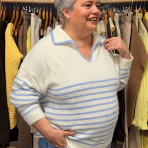 Femme ronde souriante aux cheveux courts gris portant un pull blanc à fines rayures bleues en maille douce, col polo contrasté bleu, manches longues posant dans une boutique de vêtements grande taille avec portants de pulls jaunes et beiges en arrière-plan.