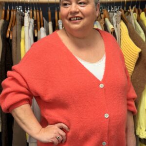 Femme ronde portant un gilet corail boutonné sur le devant, posant dans une boutique de vêtements grande taille avec portants de pantalons et pulls en arrière‑plan.