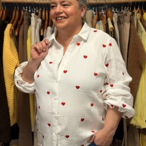 Femme ronde aux cheveux courts gris portant une chemise blanche en gaze de coton à motifs cœurs rouges brodés, col chemise à manches longues bouffantes, posant souriante dans une boutique de vêtements grande taille avec portants de pulls jaunes et pantalons en arrière-plan.