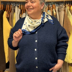 Femme ronde aux cheveux courts gris portant un gilet bleu marine en maille douce à boutons, foulard blanc à motifs cœurs bleus noué autour du cou, posant souriante dans une boutique de vêtements grande taille avec portants de pulls jaunes et pantalons en arrière-plan.