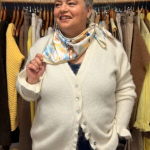 Femme ronde aux cheveux courts gris portant un gilet écru en maille douce à boutons, foulard blanc à motifs colorés noué autour du cou, posant souriante dans une boutique de vêtements grande taille avec portants de pulls jaunes et pantalons en arrière-plan.