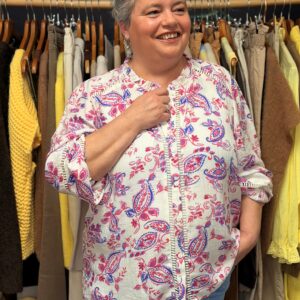 Femme ronde aux cheveux courts gris portant une tunique oversize blanche en gaze de coton à motifs cachemire violets et roses, col V à manches longues bouffantes froncées, posant souriante dans une boutique de vêtements grande taille avec portants de pulls jaunes et pantalons en arrière-plan.