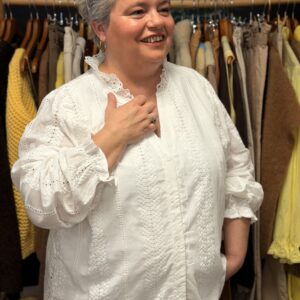 Femme ronde aux cheveux courts gris portant une chemise blanche en broderie anglaise à dentelle, col V festonné à manches 3/4 bouffantes, posant souriante dans une boutique de vêtements grande taille avec portants de pulls jaunes et pantalons en arrière-plan.