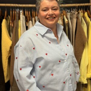 Femme ronde aux cheveux courts gris portant une chemise bleu clair à fines rayures et motifs cœurs rouges brodés, col chemise, manches longues larges, posant souriante dans une boutique de vêtements grande taille avec portants de pulls jaunes et beiges en arrière-plan.
