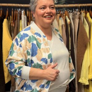 Femme ronde aux cheveux courts gris portant un bomber léger imprimé feuilles ginkgo bleu, vert et beige sur fond clair, associé à un top irisé écru, posant de face dans une boutique de vêtements.