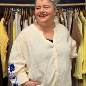 Femme ronde aux cheveux courts gris portant une blouse oversize en gaze de coton beige clair, col tunisien boutonné et manches 3/4 amples avec broderies florales bleues sur le bas de manche, posant dans une boutique de vêtements