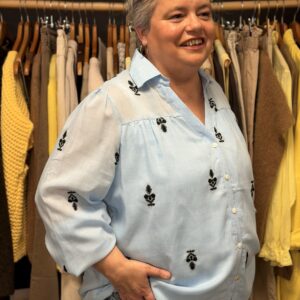 Femme ronde aux cheveux courts gris portant une blouse en gaze de coton bleu ciel, col V avec petit col chemise, manches 3/4 amples et motifs brodés noirs, posant dans une boutique de vêtements.
