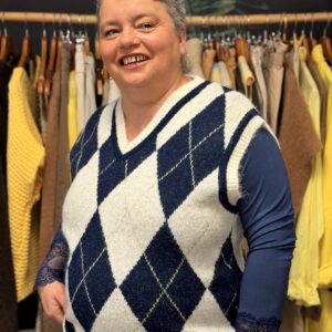 Femme ronde aux cheveux courts gris portant un pull sans manches à losanges bleu marine et écru, col V, superposé à un haut manches longues bleu marine, posant dans une boutique de vêtements