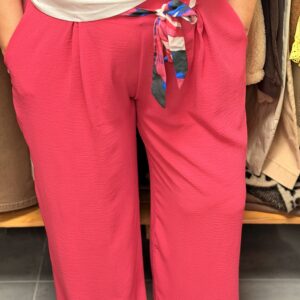 Pantalon fluide taille haute rose fuchsia plissé structuré en tissu fluide gaufré, ceinture large nouée foulard multicolore rayé, coupe évasée fluide aux chevilles, porté dans une boutique avec portants vêtements en arrière-plan.