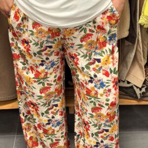 Pantalon fluide imprimé fleurs multicolores orange, rose, vert et bleu sur fond blanc, coupe fluide évasée aux chevilles, porté dans une boutique de vêtements.