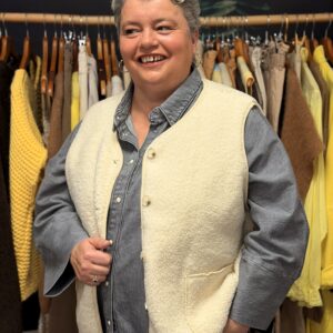 Femme ronde aux cheveux courts gris portant un gilet en maille bouclette beige clair, col chemise à boutons, superposé à une chemise bleu marine, posant souriante dans une boutique de vêtements.