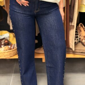 Pantalon jean bleu brut, coupe droite légèrement évasée, taille haute sans poches avant apparentes, détail œillets dorés sur les côtés des jambes, porté dans une boutique