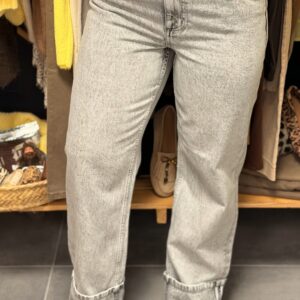 Pantalon jean gris clair effet used, coupe flare évasée aux chevilles, porté dans une boutique avec portants vêtements et paniers tressés