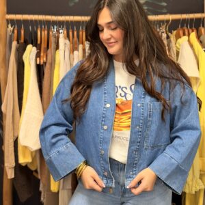 Femme aux cheveux longs bruns portant une veste en jean bleu à boutons métalliques, col chemise, manches larges.