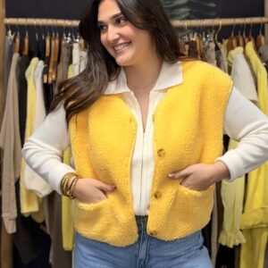 Femme aux cheveux longs bruns portant une veste sans manches jaune, effet moutonné, mains dans les poches plaquées.