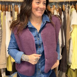 Femme aux cheveux longs bruns ondulés de dos, portant un gilet sans manches violet prune en maille bouclette, superposé à une chemise denim bleu.