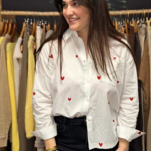 Femme portant une chemise blanche en coton à motifs cœurs rouges brodés, col chemise à manches longues, posant souriante dans une boutique de vêtements.