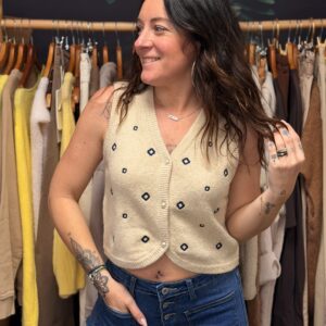 Femme brune portant un gilet sans manches en maille douce, avec broderies contrastées. Coupe courte, 3 boutons et encolure en V.