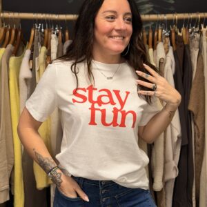 Femme sourainte portant un tee-shirt blanc à coupe droite, orné d’un message rouge « Stay Fun » sur la poitrine.
