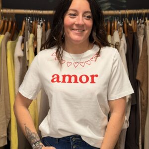 Femme aux cheveux longs bruns portant un t-shirt blanc à inscription rouge “amor” et col décoré de petits cœurs, associé à un jean bleu taille haute boutonné, posant dans une boutique de vêtements.