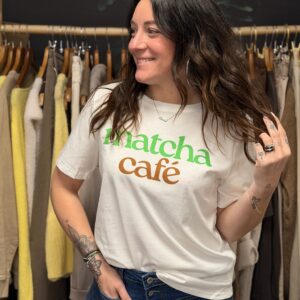 Femme aux cheveux longs bruns portant un t-shirt blanc à message “matcha café” écrit en vert et orange, manches courtes, associé à un jean bleu taille haute boutonné.